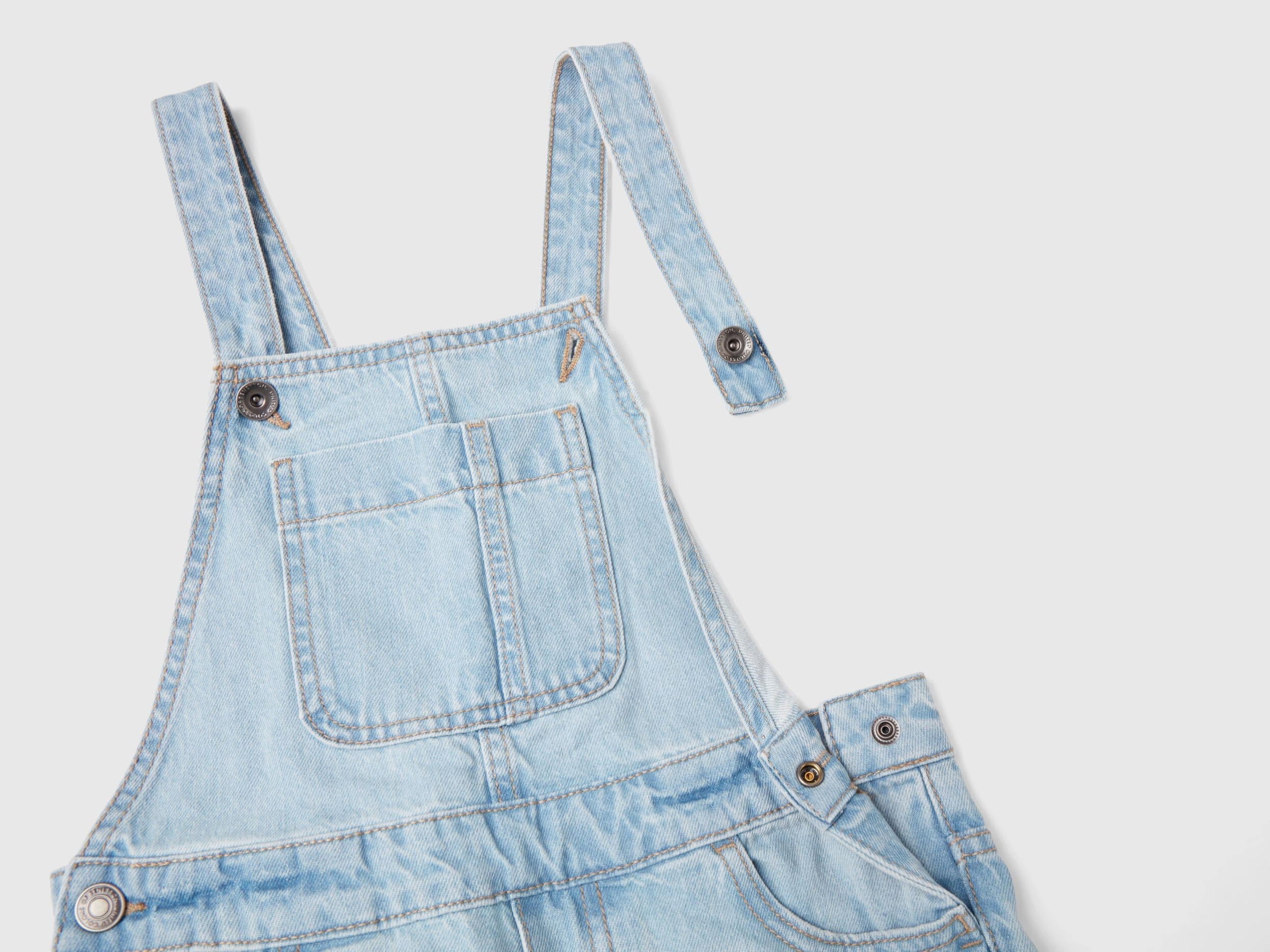 Close-up of light wash denim dungaree shorts for girls in Kuwait, showing strap and button detail.