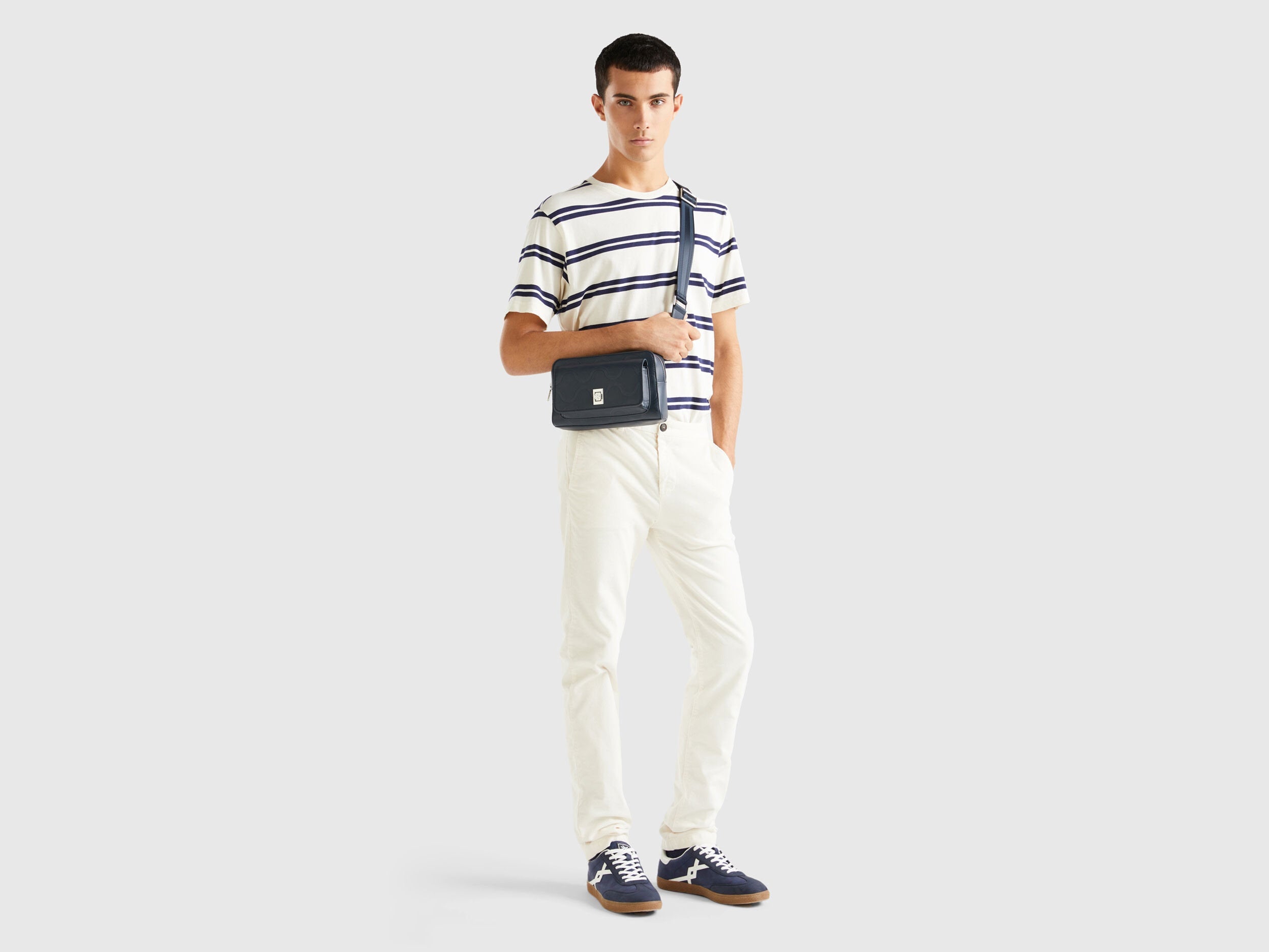 A male model wearing white stretch velvet trousers, paired with a striped shirt and bag, showcasing velvet jeans for men in Kuwait for a stylish look.