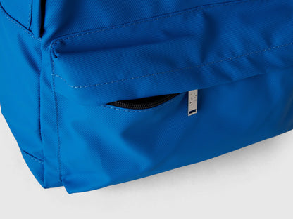 Close-up detail of a blue nylon backpack, focusing on a zippered side pocket and a small white brand tag.