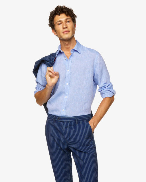 Male model wearing a light blue long-sleeve linen shirt, with a dark blue striped jacket draped over his shoulder, showcasing a casual look perfect for linen shirts in Kuwait.